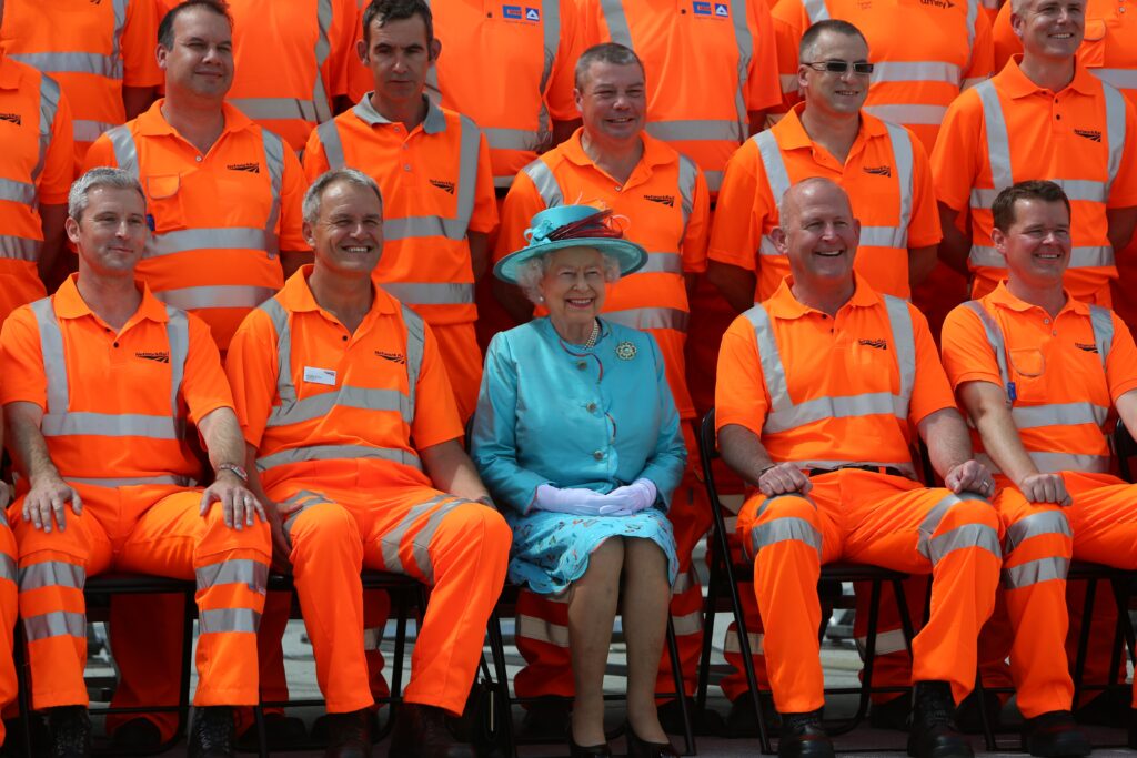 Her Majesty The Queen with the people that rebuilt Reading Station. The Queen is dressed in blue, with the surrounding engineers wearing orange high visibility clothing.